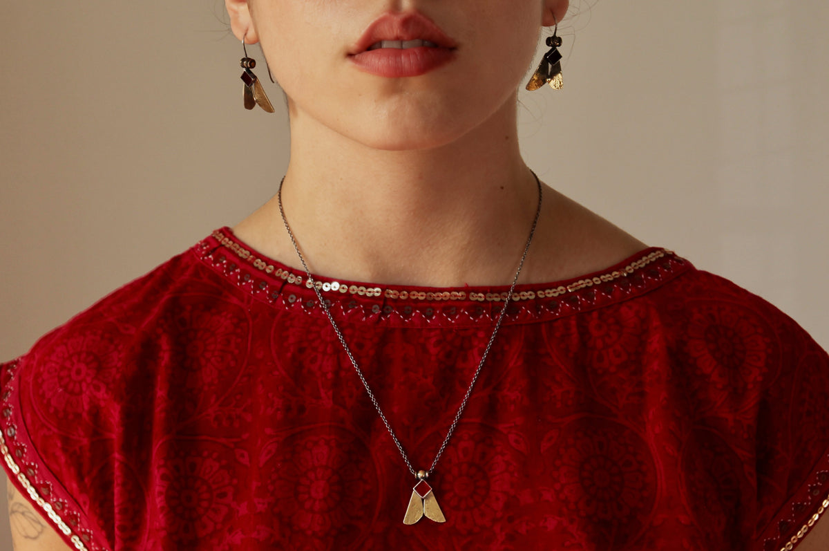 A close-up of a person wearing a silver-toned necklace with a gold-toned triangular pendant and matching dangling earrings, paired with a red embroidered top against a plain beige background.