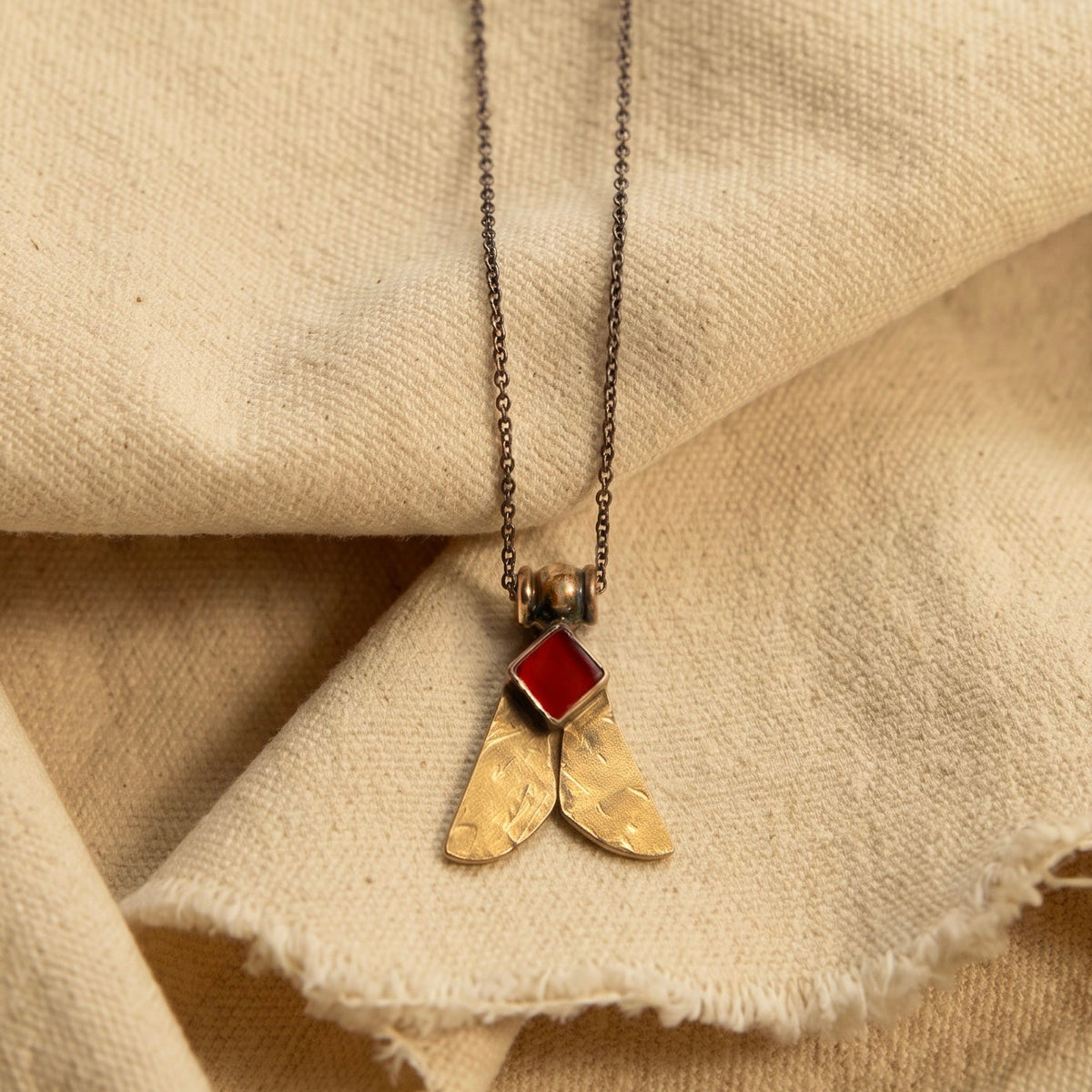 A close-up of a necklace with a textured gold pendant shaped like two wings, topped with a small red gemstone, resting on draped beige linen fabric.
