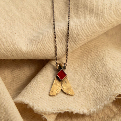 A close-up of a necklace with a textured gold pendant shaped like two wings, topped with a small red gemstone, resting on draped beige linen fabric.