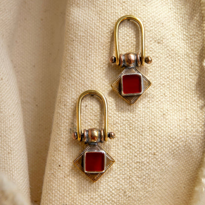 A pair of vintage-style gold and silver earrings with square red gemstones, displayed on a textured beige fabric background.