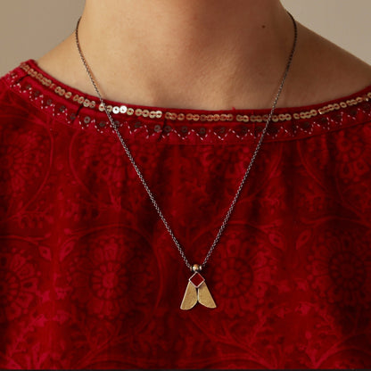 A close-up of a model wearing a textured red velvet top with gold trim, a silver necklace with a gold pendant, and matching dangling earrings against a plain beige background.